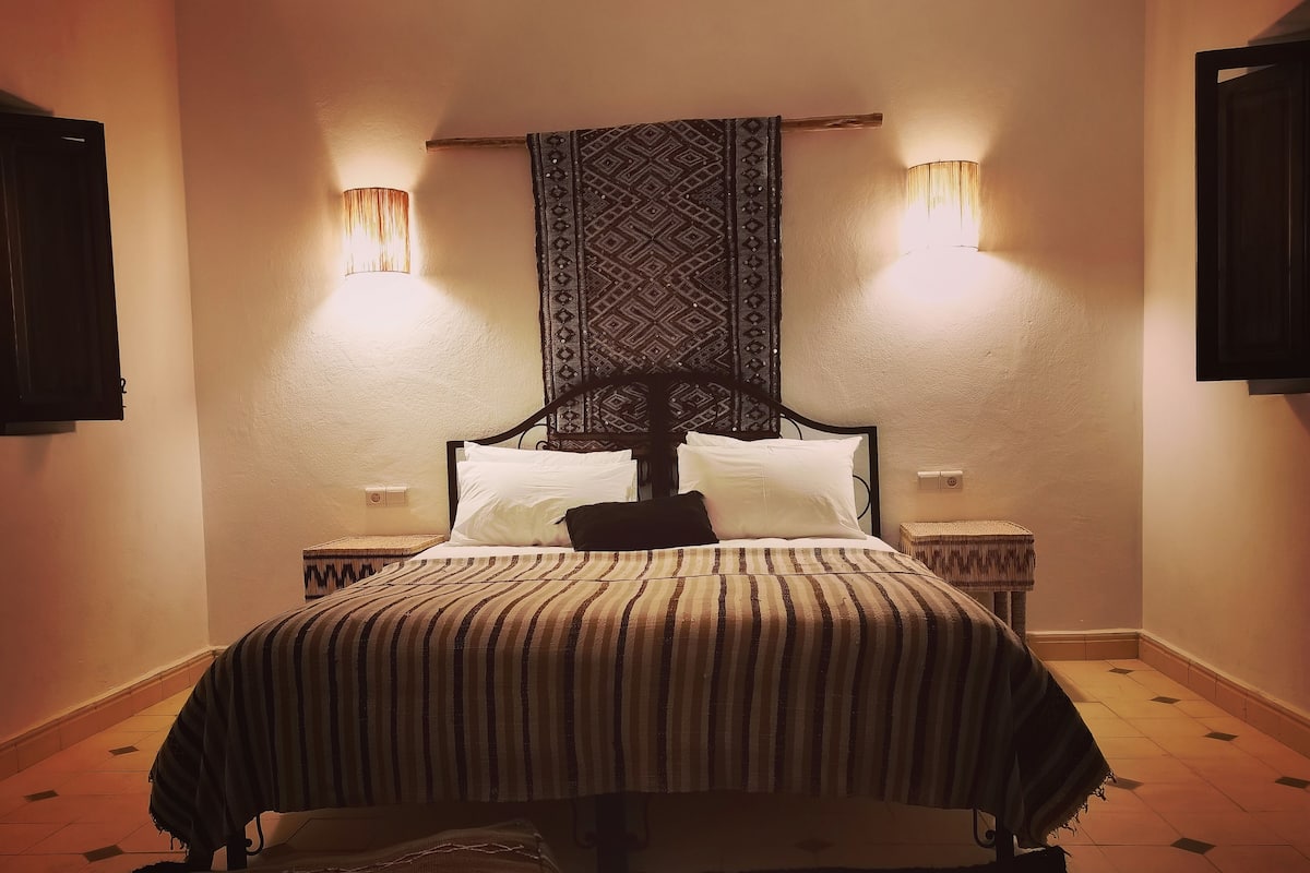 Ein Bett mit gestreifter Bettdecke, zwei Wandlampen und einem gemusterten Wandteppich über dem Kopfteil.
