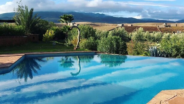 Un ciel d’un bleu limpide parsemé de quelques nuages, une piscine qui reflète le ciel et, au loin, un paysage montagneux.