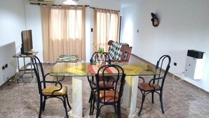 A dining area with a glass table surrounded by four chairs, a television on a stand, and a plant on the table.