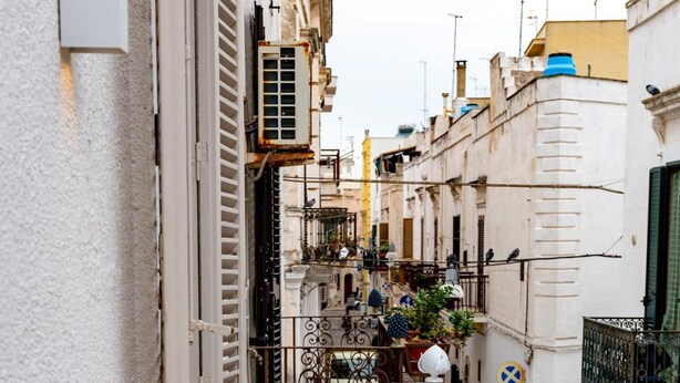 A narrow urban street with buildings on either side, a white balcony with ornate metal railings, and a small table with a potted plant. - D'aprile Dimore (Polignano a Mare)