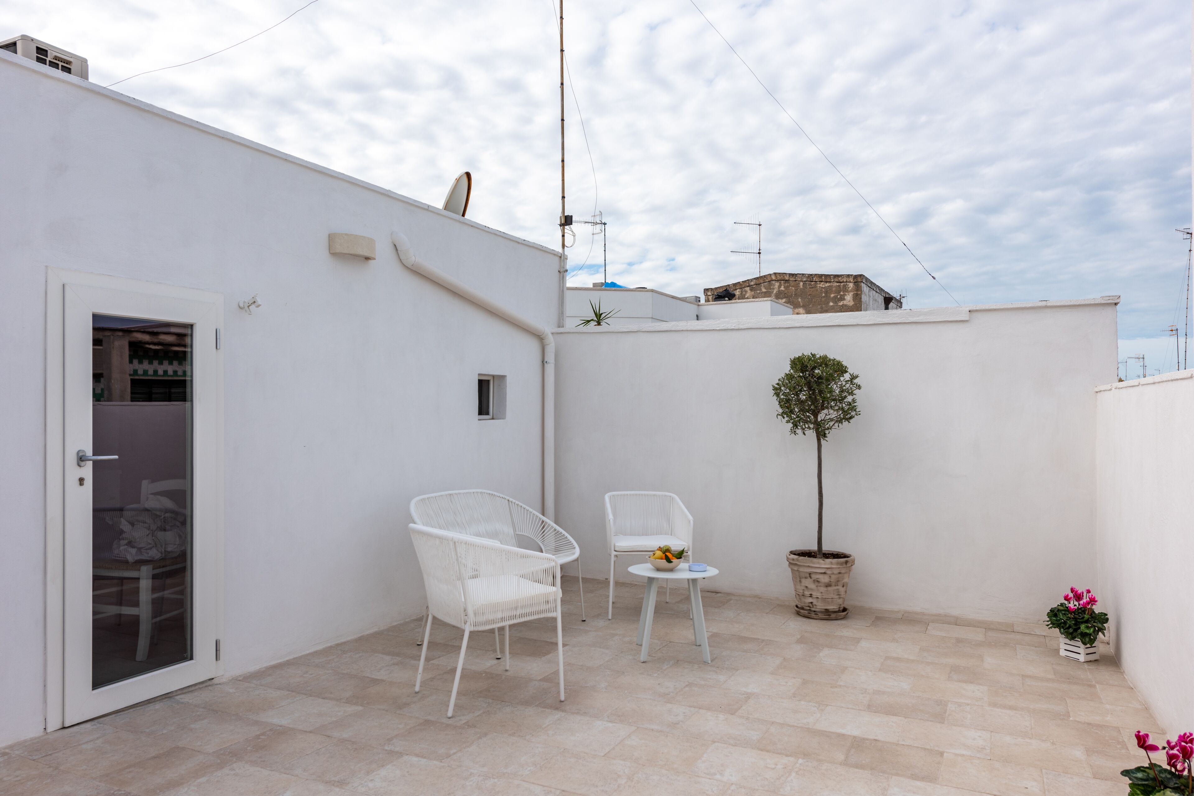 Una terraza en la azotea con dos sillas de mimbre blancas, una mesa redonda pequeña con un cuenco de fruta, un árbol en maceta y una puerta con panel de vidrio.