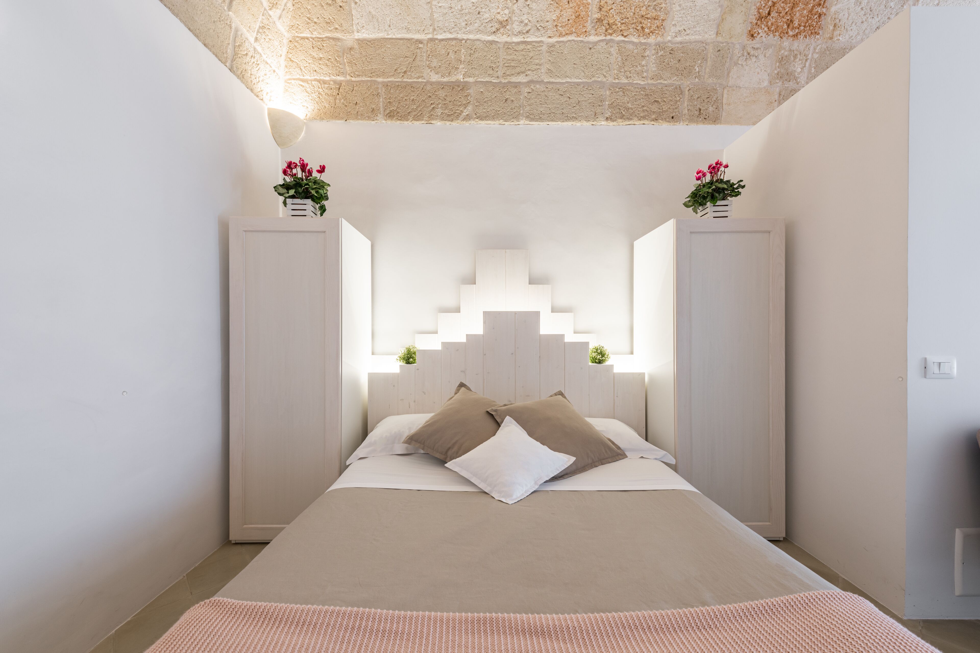A bedroom with a bed centered between two white cabinets, a stone ceiling, and a small plant on each cabinet.