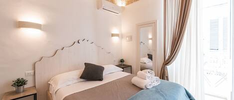 A hotel room with a bed, bedside tables, a potted plant, and a view of the outside.