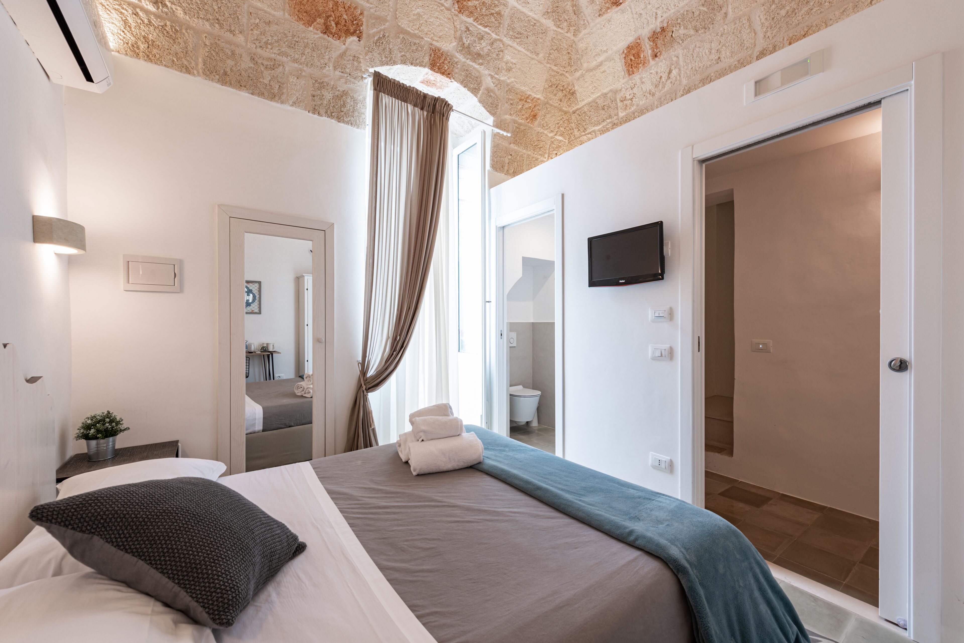 A modern hotel room with a brick ceiling, a large bed with a blue blanket, a flat-screen TV, and a bathroom visible through an open door.