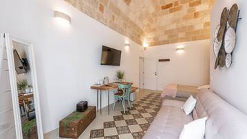 Uma sala de estar acolhedora, com piso xadrez, um sofá, uma mesa de jantar com cadeiras, uma televisão na parede e um espelho refletindo o ambiente.