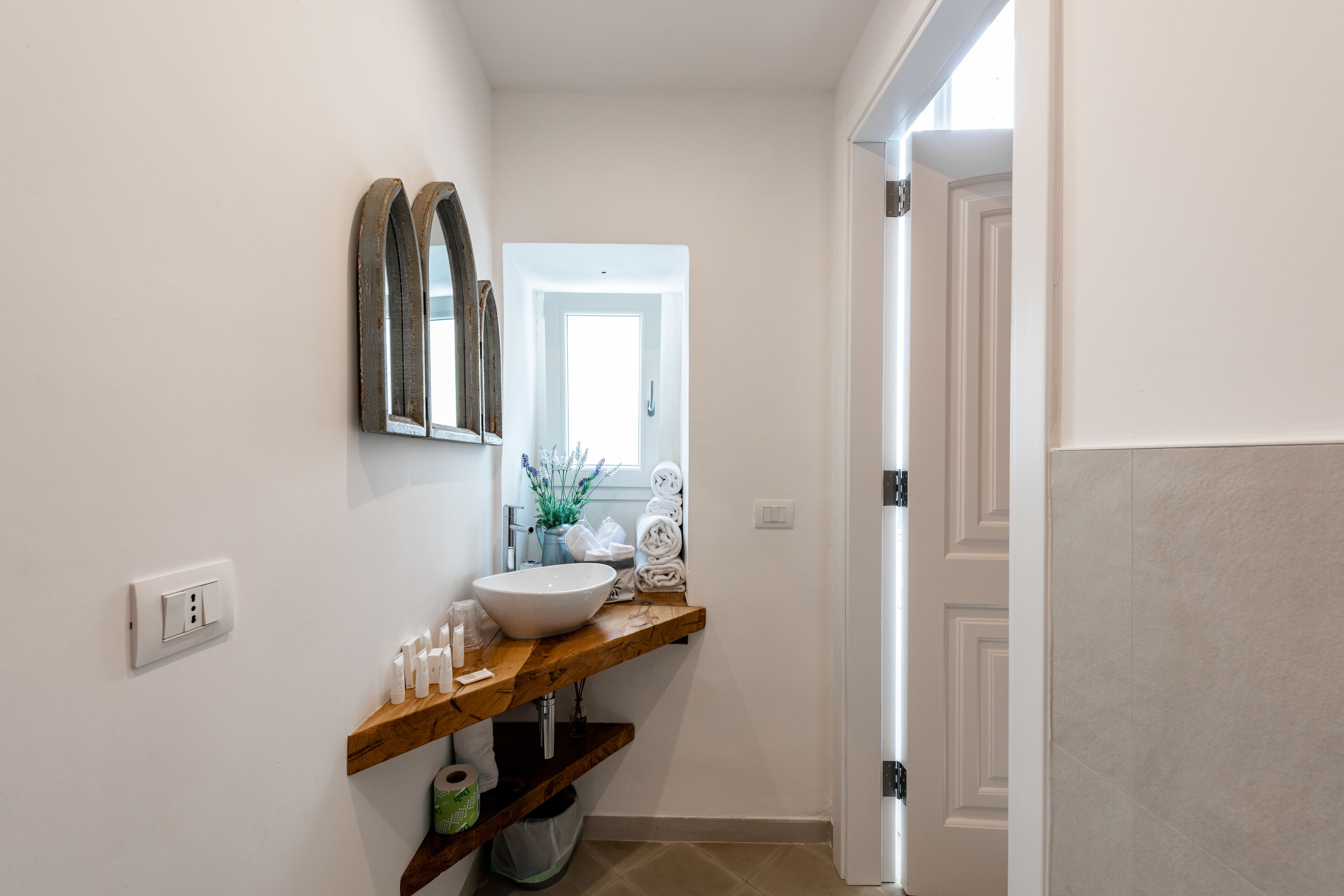 A bathroom with a wooden vanity, a white sink, and a mirror. There is a towel rack with folded towels and a small plant on the vanity.