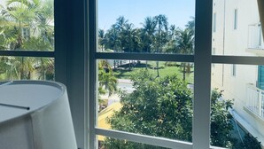 A room with a large window offering a view of a well-maintained garden with palm trees and a building in the background. - Ocean view, luxury STUDIO, JACUZZI on Ocean drive (Miami Beach)