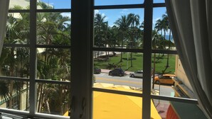 A view through a window with a view of palm trees, a grassy area, and a road with cars. - Ocean view, luxury STUDIO, JACUZZI on Ocean drive (Miami Beach)