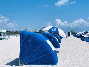Una playa con hileras de sombrillas azules y sillas de playa, una torre de salvavidas y edificios al fondo.