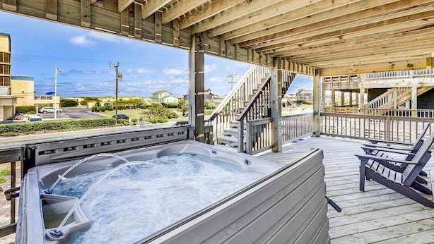 Een houten terras met een jacuzzi, buitenmeubels en een trap naar een hoger gelegen niveau.