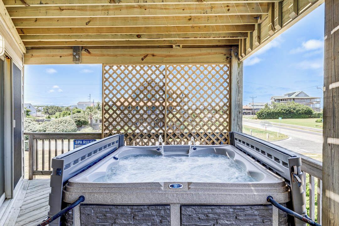 Une terrasse en bois avec un jacuzzi installé sous une pergola, un panneau indiquant les règles du spa, et une vue sur une route et des bâtiments en arrière-plan.