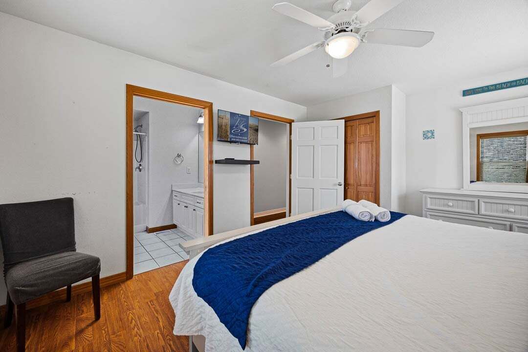 A bedroom with a bed, a chair, a ceiling fan, a shower, and a door leading to a bathroom.
