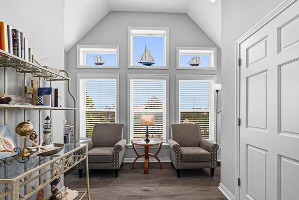 A room with a sloped ceiling, a bookshelf, two armchairs, a round table, and a window with a sailboat view.