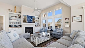 A living room with a fireplace, bookshelves, and a view of trees. - Pelican Pointe | 355 ft from the Beach | Private Pool, Hot Tub | Corolla (Corolla)