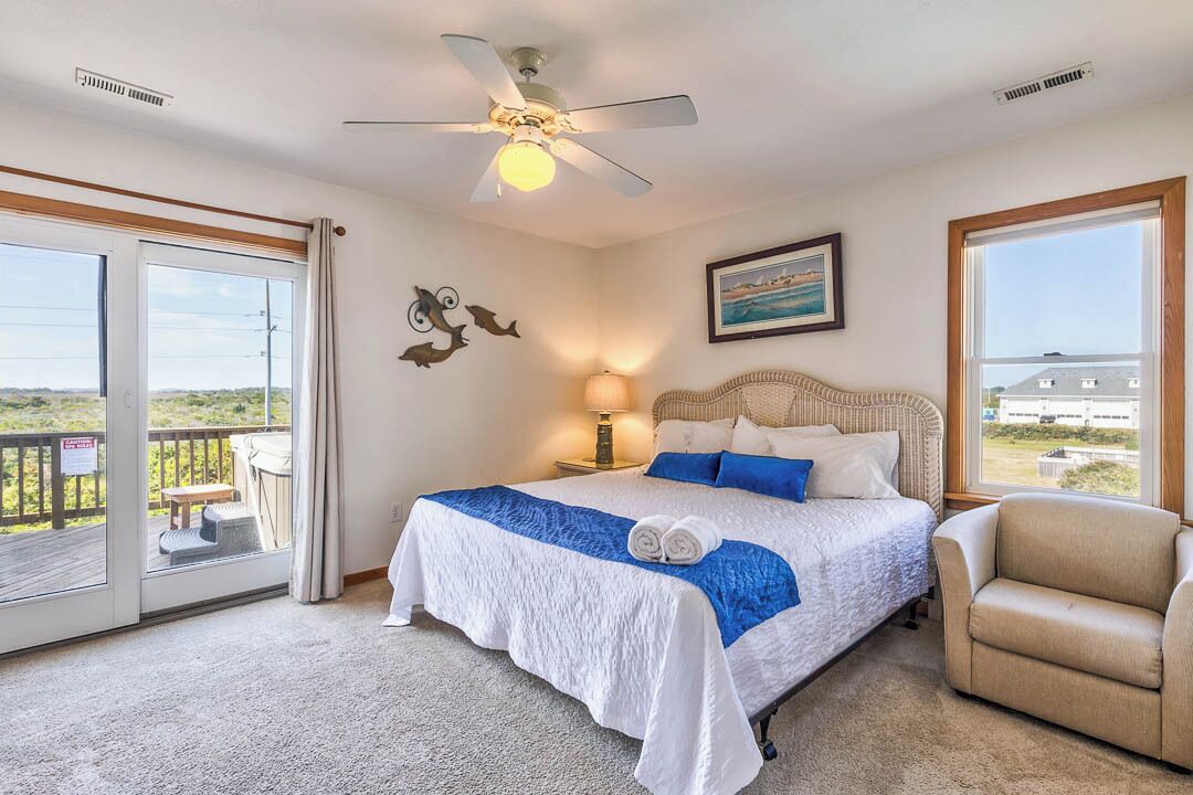 A bedroom with a bed, a ceiling fan, a nightstand with a lamp, a chair, and a large window with a view.