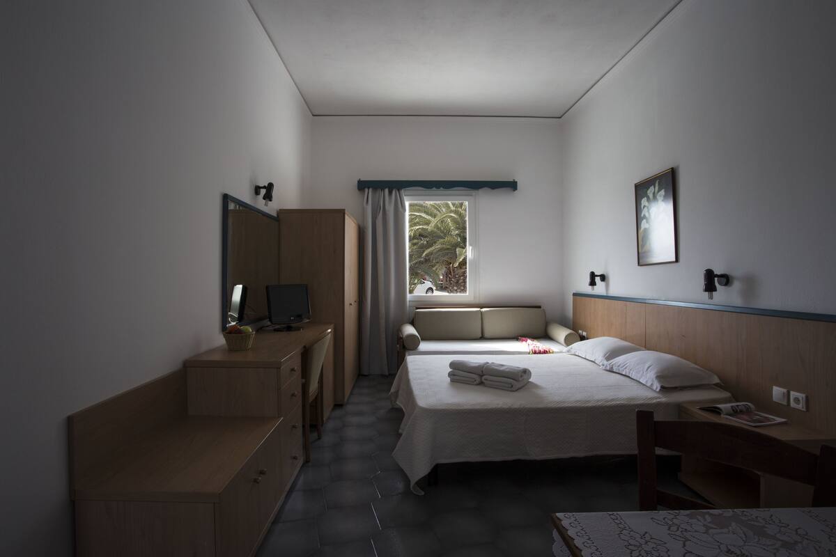 A hotel room with a bed, a desk, a chair, a wardrobe, a window with a view of palm trees, and a painting on the wall.