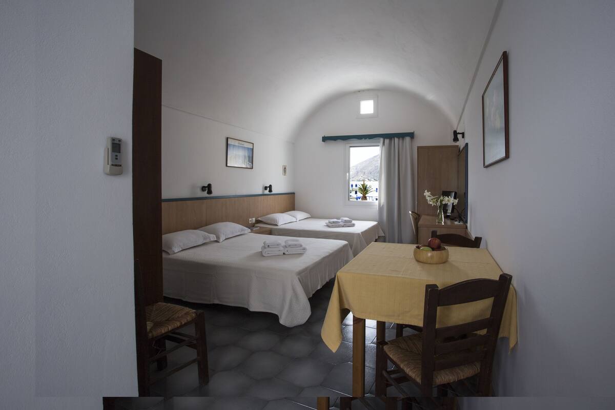 A compact hotel room with two single beds, a small dining table, and a window with curtains.