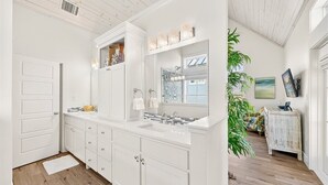 A white kitchen with a large island, wooden flooring, and a view of a living area with a plant and a painting. - Gracious Home w/ Lake Views, Fire Pit, and More Amenities in Cinnamon Shore (Port Aransas)