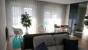 A modern living room with a grey sofa, two dark cushions, a potted plant, and a dining area with a table and chairs.