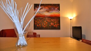 A clear glass vase with white sticks on a wooden table, a painting of a sunset on the wall, and a lamp to the right. - Street Art House (Catania)