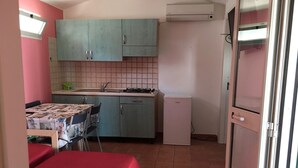 Cocina compacta con armarios color turquesa, un refrigerador pequeño y un comedor con mesa y sillas rojas.