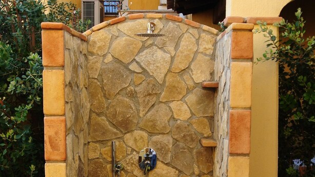 A stone shower enclosure with a metal faucet and a handheld showerhead, surrounded by a brick wall and a yellow building.