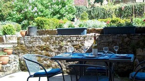 An outdoor dining area with a stone wall, metal chairs, and a table set for two with wine glasses and a bottle. - Landmarked stone house by a stream with extraordinary charm (Frontenay)