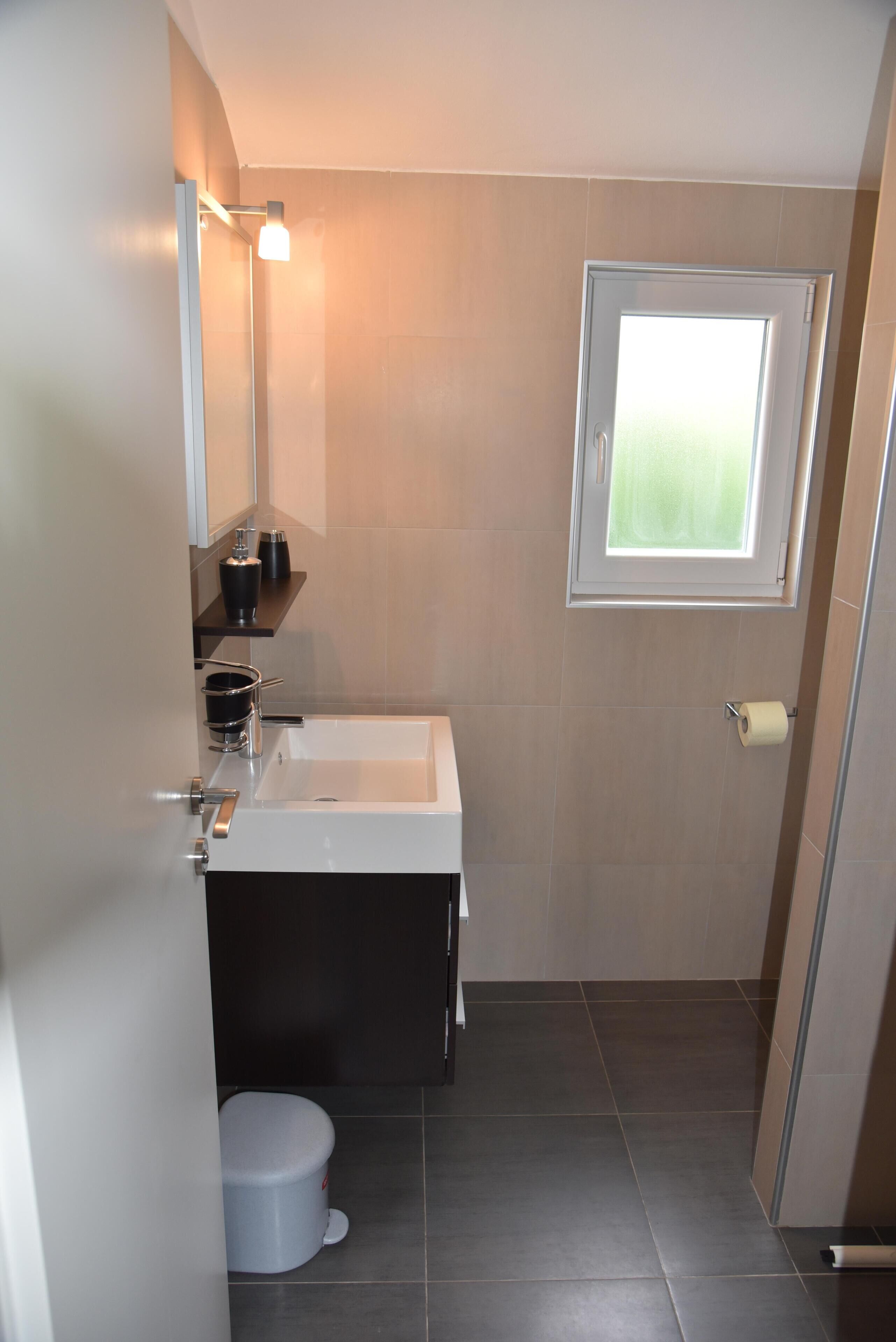 A compact bathroom with a white sink, a mirror, a toilet, and a window.