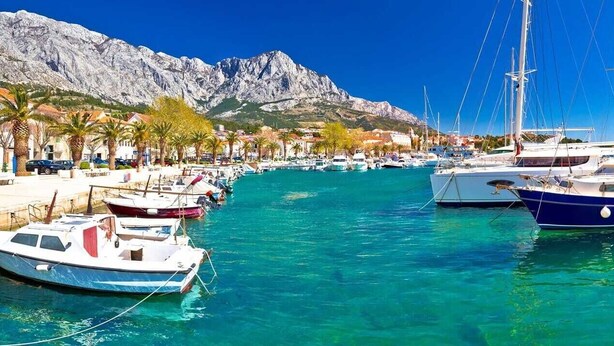 A marina with boats docked, a waterfront promenade, and a mountainous backdrop.