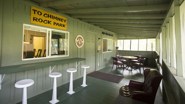 Eine überdachte Veranda mit einem Schild, das den Weg zum Chimney Rock Park weist. Weiße Hocker, ein Tisch mit Stühlen und eine Bank mit Kissen sind vorhanden.