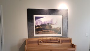 A wooden piano with a bench in a room with a white wall and a framed painting above it.