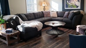 A living room with a sectional sofa, a round coffee table, a side table with books, a lamp, and a framed picture on the wall.
