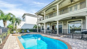 En swimmingpool med brobelagt terrasse, en overdækket terrasse med udendørs møbler og en toetagers bygning med altaner.