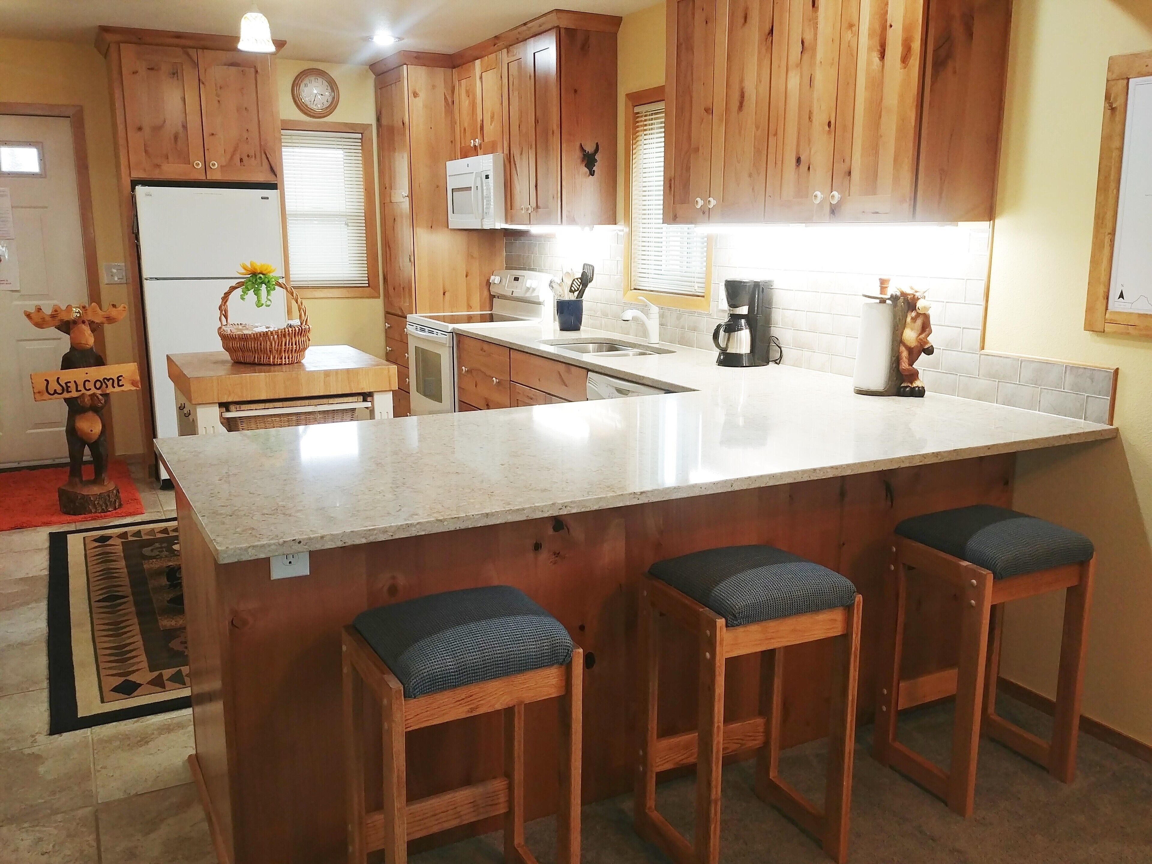A kitchen with wooden cabinets, a white refrigerator, a microwave, a coffee maker, and a sink. There are three bar stools with cushions.