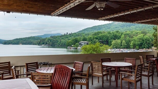 A covered outdoor seating area with tables and chairs, overlooking a lake and forested hills.