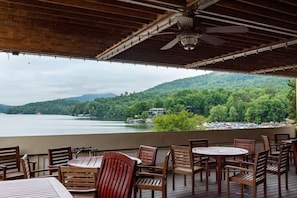 A covered outdoor seating area with tables and chairs, overlooking a lake and forested hills. - Lake Lure North Carolina | Family Lake Retreat | Pool + Lake | 2BR (Lake Lure)