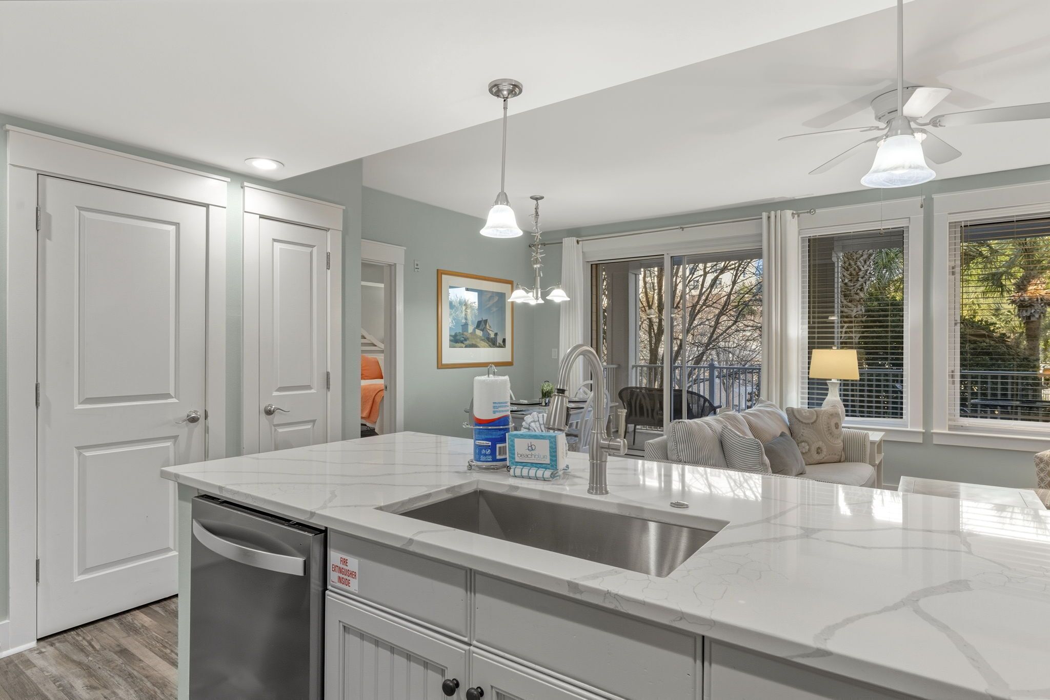 A modern kitchen with a white countertop, stainless steel sink, and a dishwasher. There are two pendant lights, a ceiling fan, and a window with blinds.