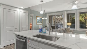 A modern kitchen with a white countertop, stainless steel sink, and a dishwasher. There are two pendant lights, a ceiling fan, and a window with blinds. - Location! Baytowne Wharf 1BR Unit w/ Balcony, Pool, Hot Tub, Tram, Beach, Gym (Miramar Beach)