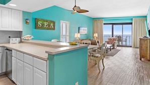 A kitchen with a large island, white cabinets, and a ceiling fan. There is a dining area with a table and chairs, and a view of the ocean through the windows.