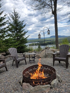A fire pit with chairs and a table, surrounded by rocks, with a view of a lake and mountains. - Spectacular Panoramic Views in a Private Setting with ADK 5 Star Accomodations (Old Forge)