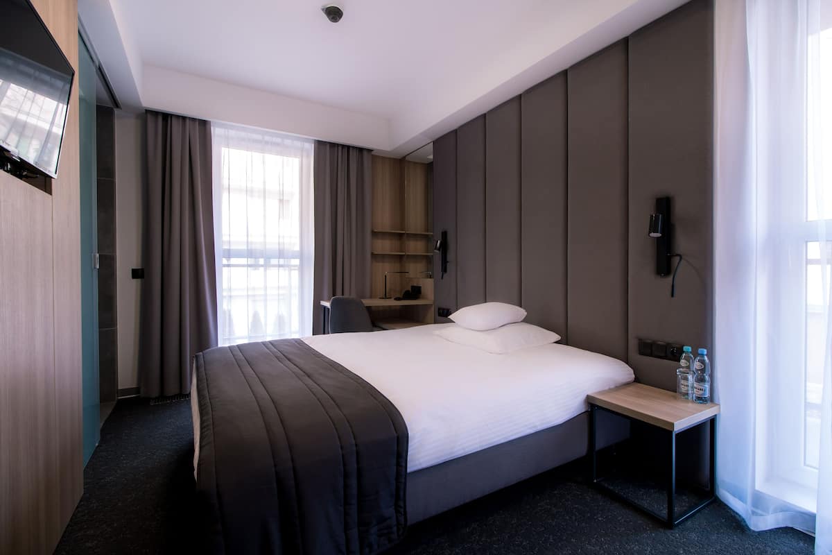 Una habitación de hotel moderna con una cama grande, un escritorio y una silla. Hay un televisor montado en la pared y una ventana con cortinas.