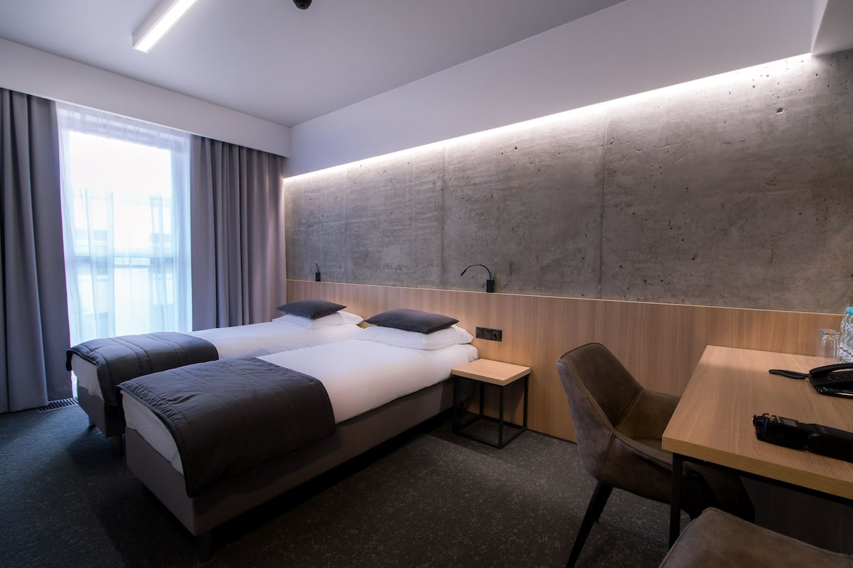 Habitación de hotel con dos camas, una mesita pequeña, una silla y un ventanal grande con cortinas.