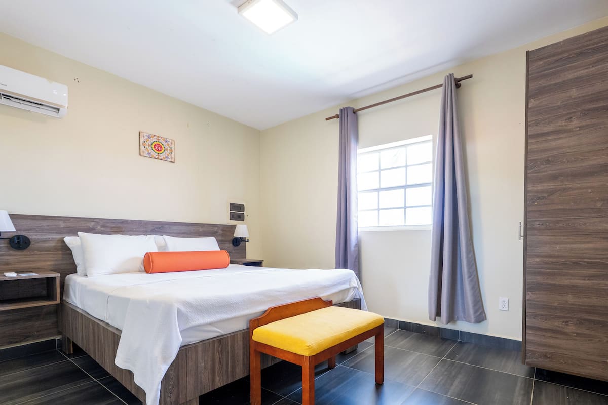 Camera d'albergo con un letto, una panca, un comodino, un condizionatore, una finestra con tende e un armadio in legno.