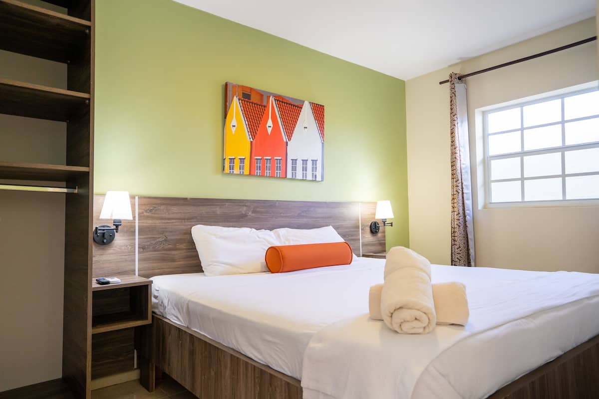Una camera d'albergo con un letto, un comodino, una lampada a parete, un quadro e una finestra con tende.