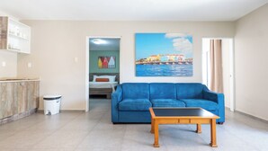 A living room with a blue sofa, a wooden coffee table, a painting of a coastal cityscape, and a kitchenette area. - Delynne Boutique Hotel (Willemstad)