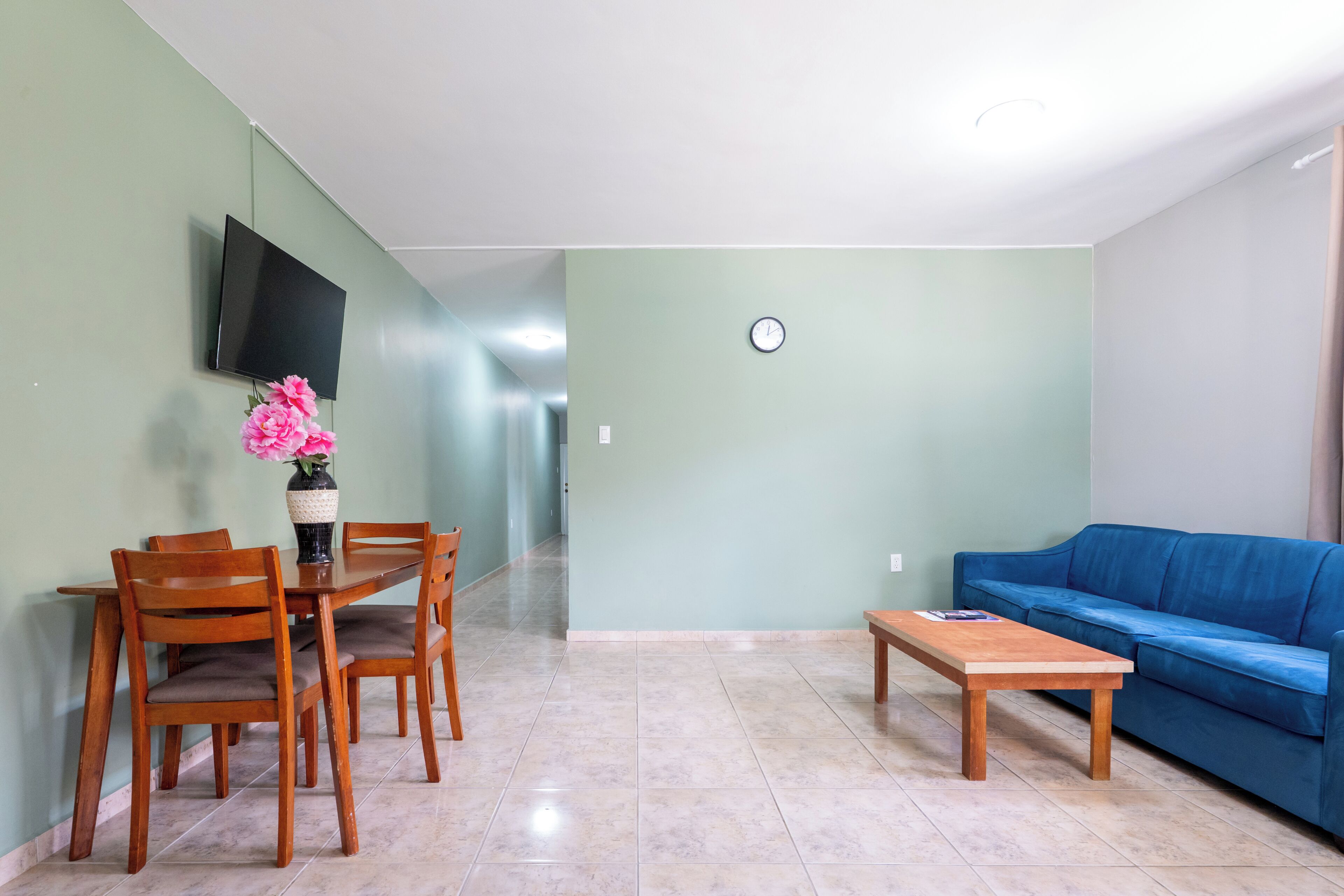 A living room with a dining table, chairs, a blue sofa, a TV mounted on the wall, and a clock on the wall.