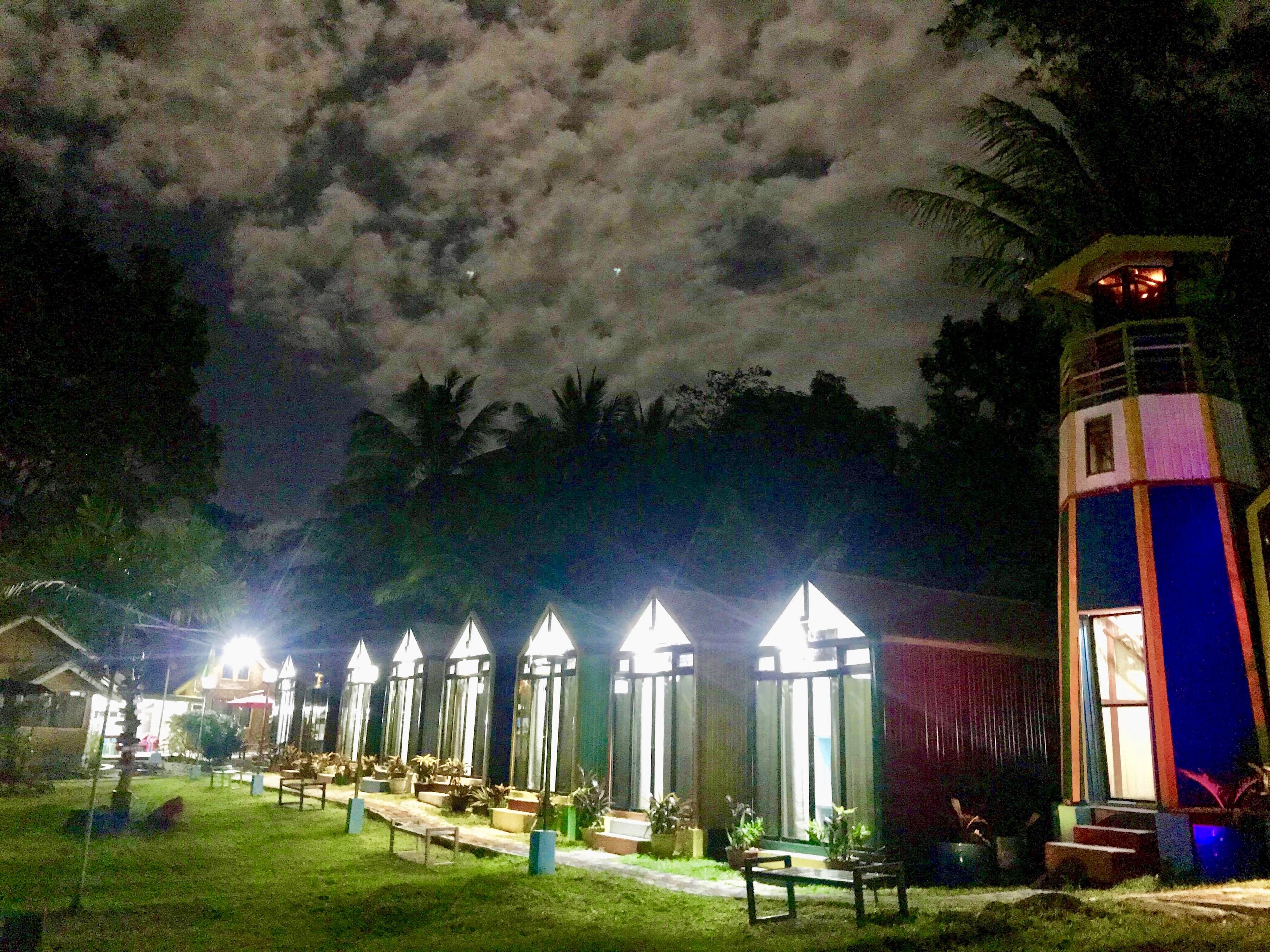 A row of brightly lit, triangular-shaped structures, possibly a hotel or resort, with a grassy area and benches in front.