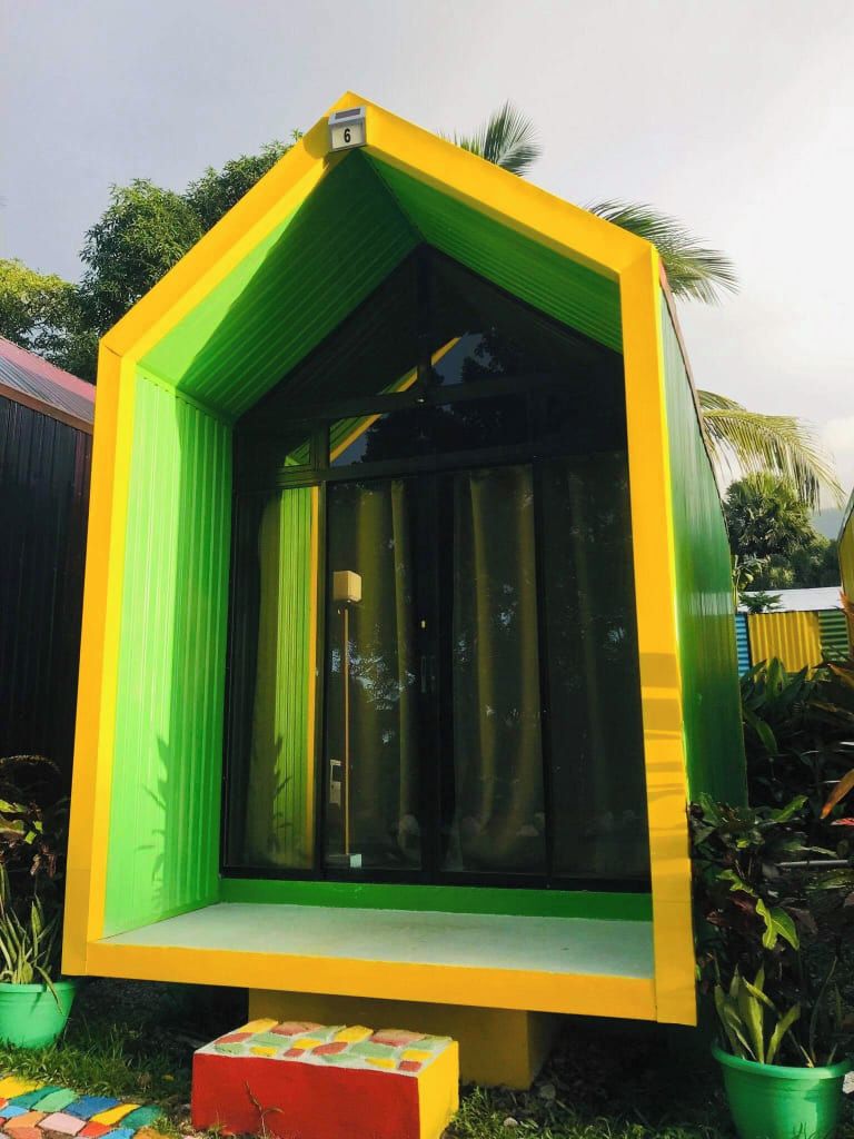 Een kleurrijke, driehoekige constructie met een raam en een deur, omgeven door groen.