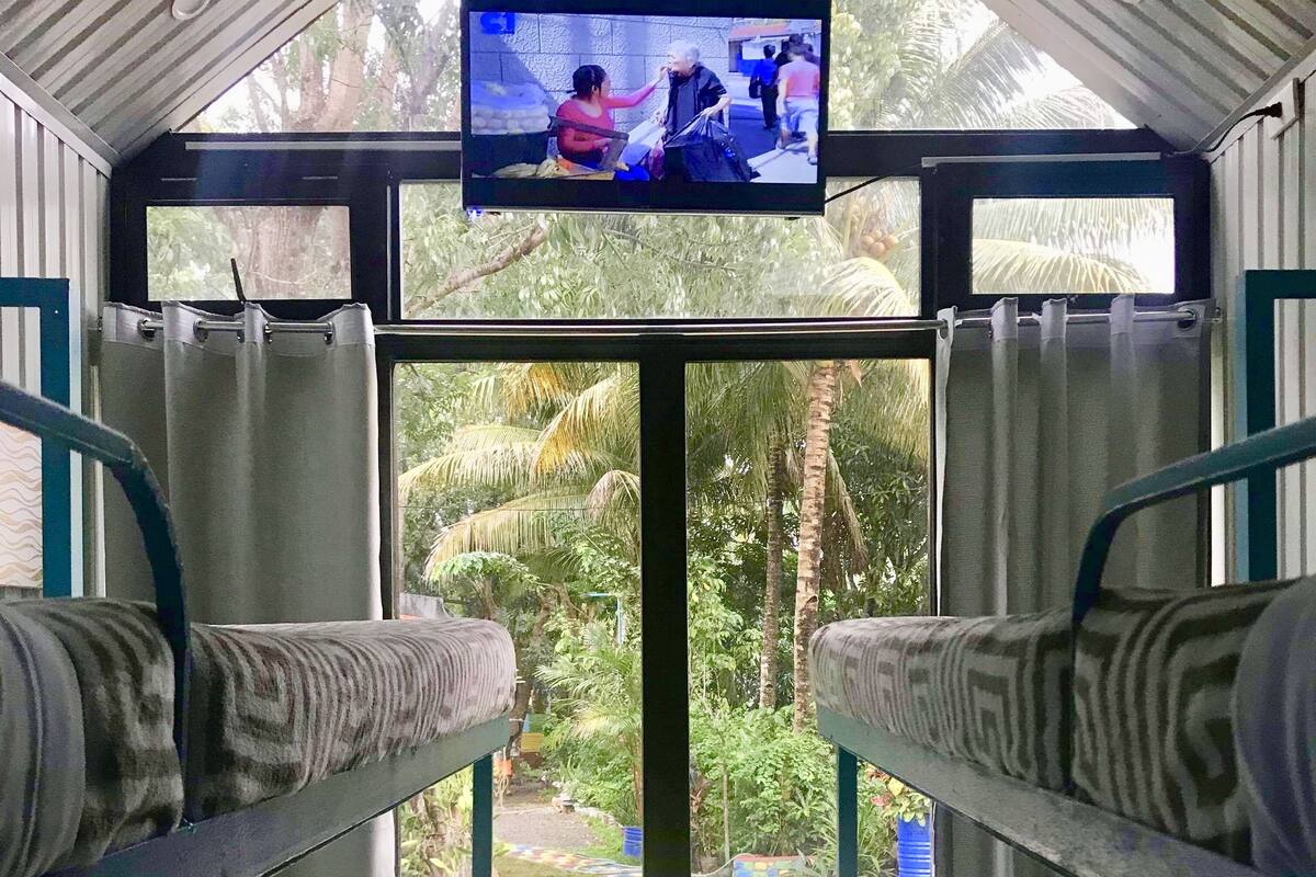 Una habitación con una televisión instalada en el techo, dos literas con cojines estampados, una mesita con botellas de agua y vista a un jardín con un sendero colorido.