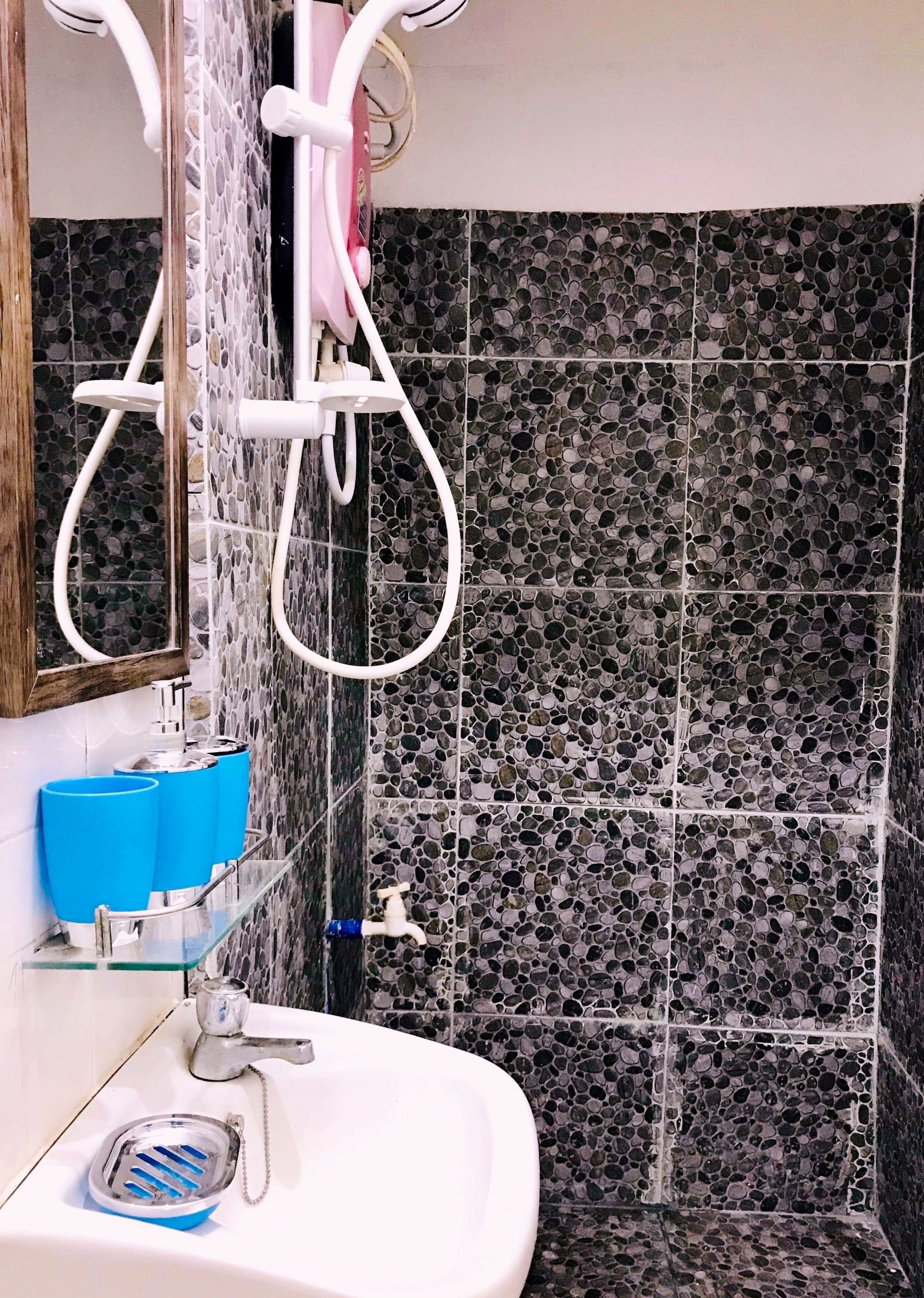 Une salle de bain avec un lavabo blanc, un miroir, une douche avec un tuyau rose et un mur carrelé orné d’un motif de petits galets.
